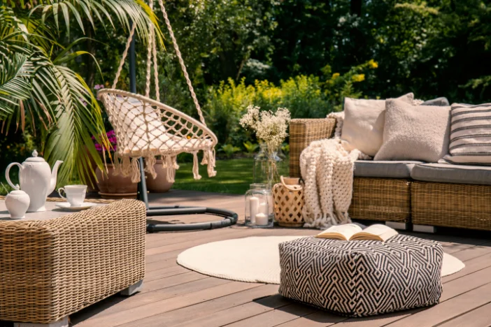 Tan wicker sofa with neutral pillows and light crocheted blanket with two-toned square pouf and round cream outdoor rug