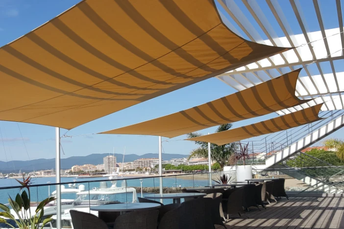 Three yellow sun shade sails suspended over outdoor dining sets at the coast