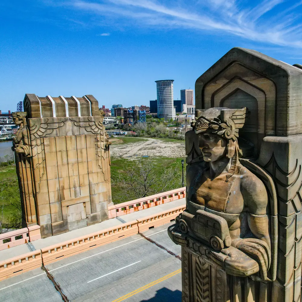 Stone carved monuments of a helmeted men holding vintage style cars on either side of an urban bridge