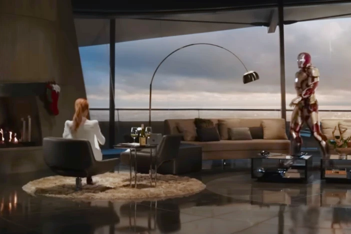 Iron Man walking toward a seated pony-tailed woman in a living room with floor-to-ceiling glass, arcing floor lamp and fireplace