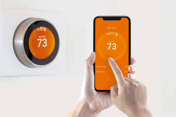 Round wall-mounted thermostat with orange face set at 73-degrees Fahrenheit by a hand-held mobile phone