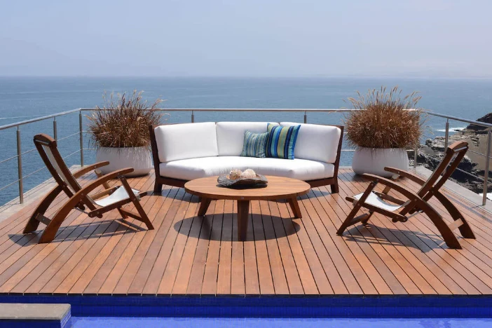 Two reddish wood deck chairs and a curved sofa with white upholstery facing a round coffee table on an oceanside deck