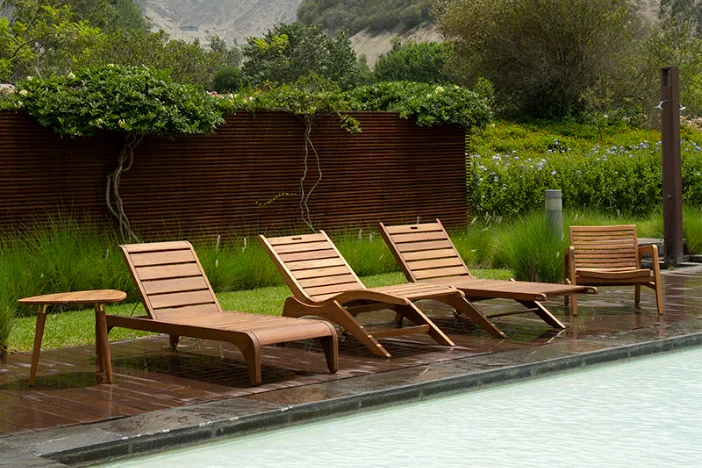 Three honey brown wooden sun loungers in different styles with an armchair and side table next to a pool with lush surroundings