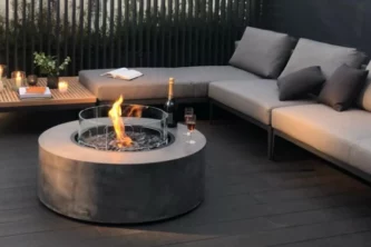 Circular dark grey composite concrete fire pit table with clear glass wind guard on a deck surrounded by a sectional