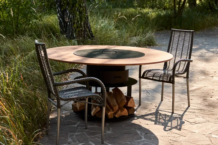 Round wood-burning fire pit table with lid and two armchairs on a backyard patio