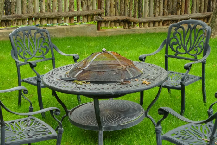 Wrought iron fire pit table with spark screen surrounded by dining armchairs on a backyard lawn