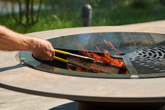 Round fire pit table with spark lid and man carefully adding firewood with tongs