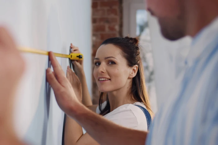 Woman and man measuring the size of a wall in an apartment using a yellow tape measure