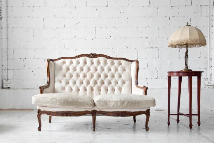 Vintage tufted white sofa with ornate wooden frame next to a tall round side table with a slender metal lamp topped by a tasseled cream shade