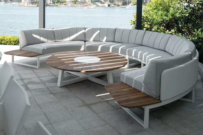 Semi-circular outdoor sectional with white metal frame, light grey angular upholstery and built-in teak side tables around a round coffee table near a body of water