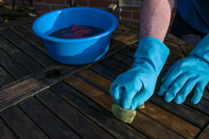 Person with blue rubber gloves cleaning between the slats of a dark patio table with water and a sponge