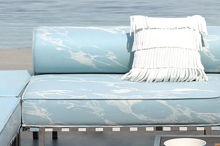 Stainless steel patio sofa with light blue acrylic upholstery with white foamy pattern and fringe pillow near a shore