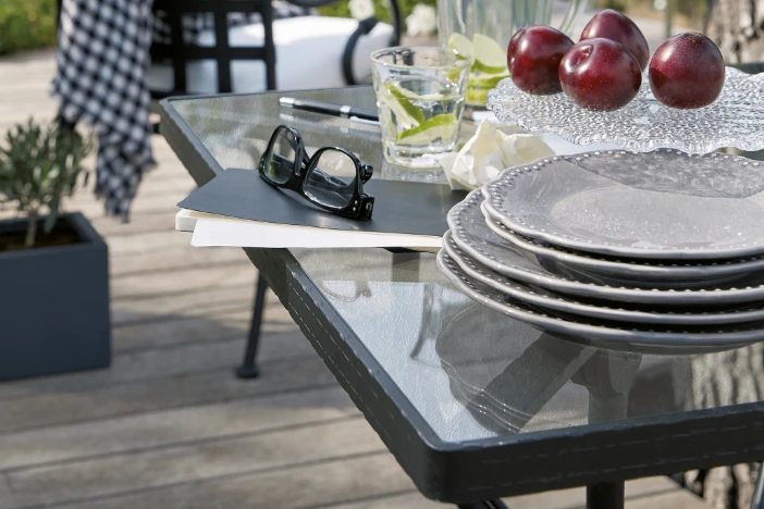 Corner of patio dining table with black metal frame, clear tempered glass top with plates, apples and reading glasses on it