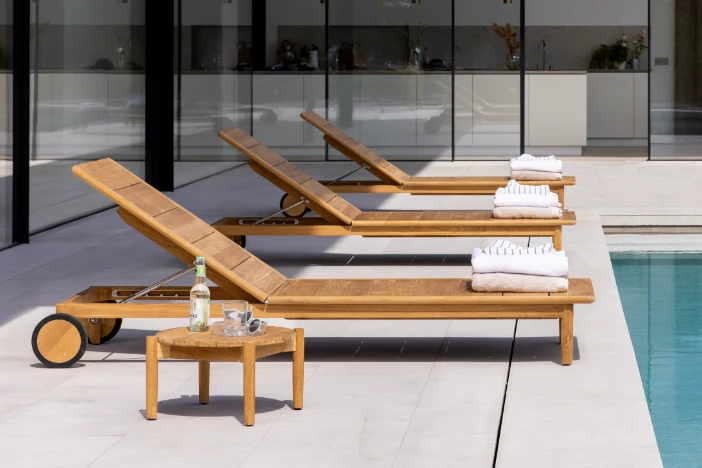 Three wooden sun loungers with wheels and matching round side table next to a pool