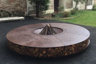 Massive flat round oxidized iron fire pit with wood stored in the side on a patio