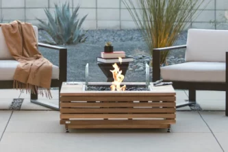 Square teak fire pit table on patio in front of two modern lounge chairs with off-white cushions and dark frames