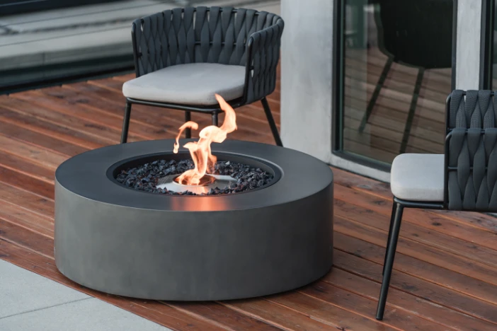 Round dark grey bio-ethanol fire pit table on hardwood deck with modern outdoor lounge chairs