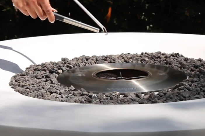 Person using a rod to manually lighting a round white fire pit with grey lava rocks surrounding the ethanol burner