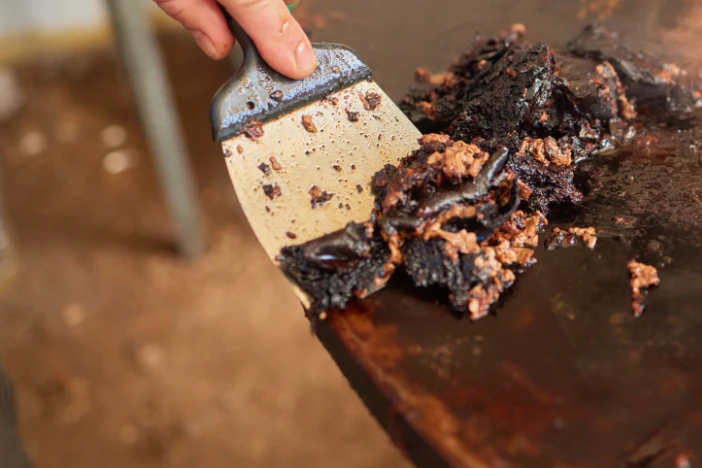 Person scraping the burnt residue off the cooking surface of a round metal fire pit