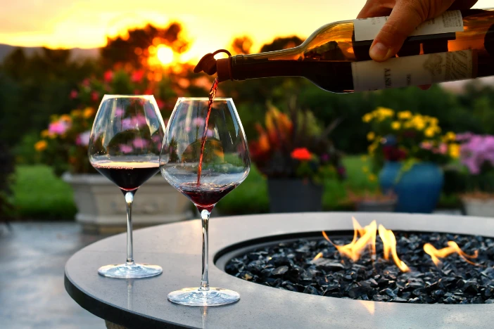 Round fire pit table with a low gas flame and person pouring red wine into two glasses at sunset