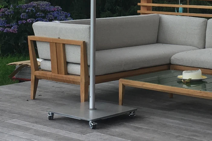 Square aluminum outdoor umbrella base with wheels on a hardwood deck near a sectional with wood frame and grey upholstery