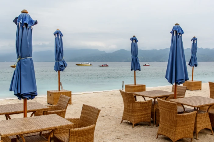 Five closed beach umbrellas with blue canopies between light brown wicker dining sets on an overcast day