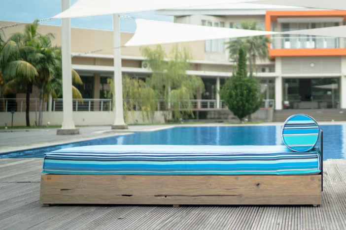 Wooden block sunlounger with blue striped seat cushion and bolster pillow with white triangular shade sails hung over a pool