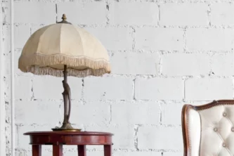 Vintage bronze lamp with umbrella-like fringed ivory shade on a reddish brown wooden side table in a white room near a tufted sofa