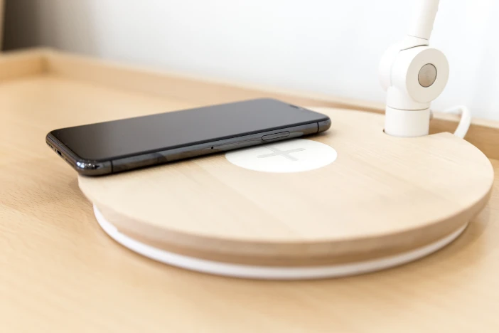 Close-up of a white table lamp with a mobile phone wirelessly charging on its round light woodgrain base