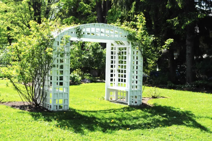 White latticed arbor with built-in benches on grass and bookended by shrubs