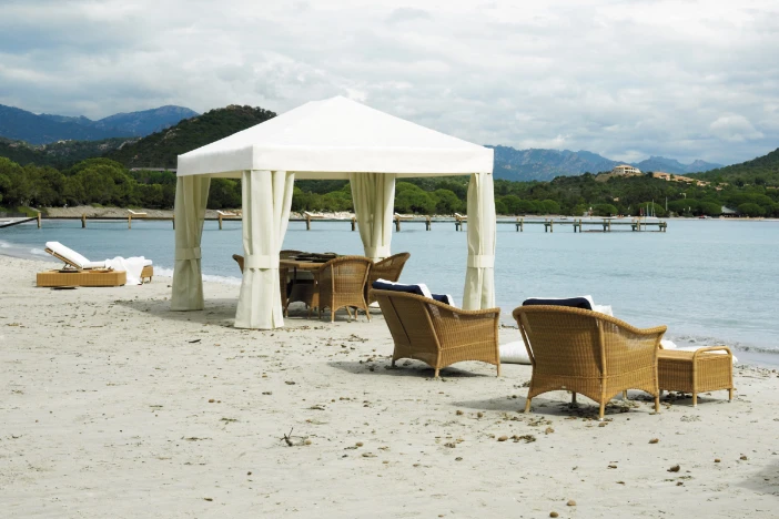 Square beach cabana with tied-off white curtains shading a wicker dining set and near matching lounge seating on an overcast day