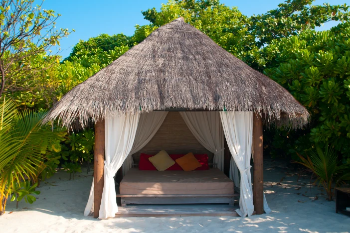 Tropical cabana with wooden pole supports, thatched roof and white curtains shading a daybed surrounded by greenery on a sunny day