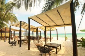 Many four post canopies with square off-white fabric roofs over lounge furniture on a beach with palm trees