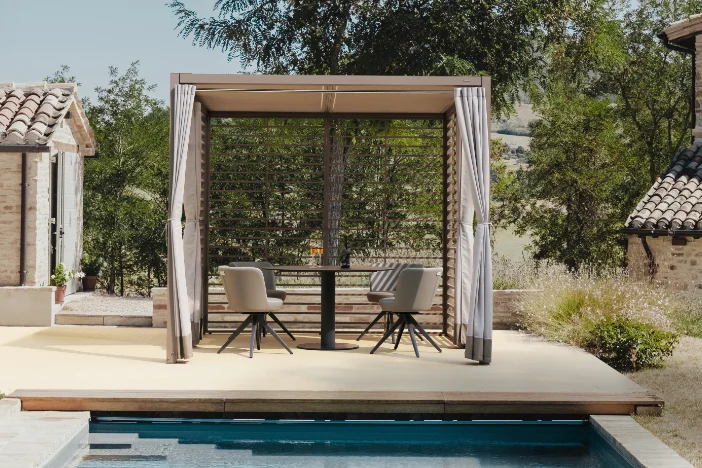 Square contemporary canopy with louvered wood sides and white curtains shading a round dining set on a poolside deck in the country