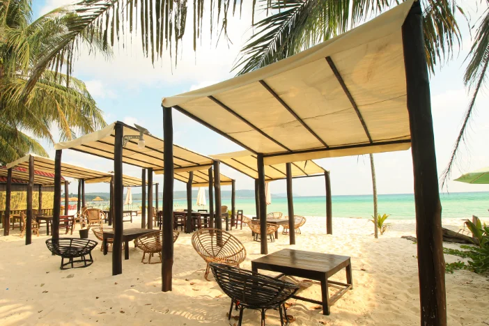 Many four post canopies with square off-white fabric roofs over lounge furniture on a beach with palm trees