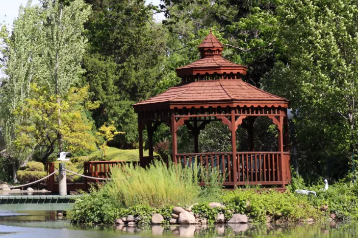 Rust-colored octagonal gazebo with three-tiered roof sitting on the edge of water in a lush green park