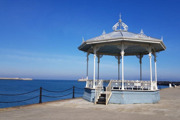 Octagonal light blue gazebo with ornate white support posts, fencing and roof accents on concrete near water on a clear day