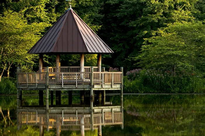 Octagonal gazebo with columns and a tall pointed dark brown roof surrounded by a wooden fence jutting into a woodside lake