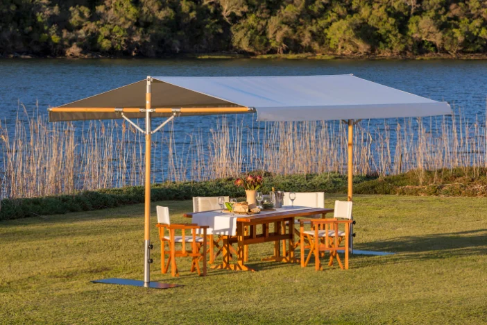 Portable rectangular pavilion with off-white fabric roof and wooden support frames shading a dining set on a lawn next to a lake