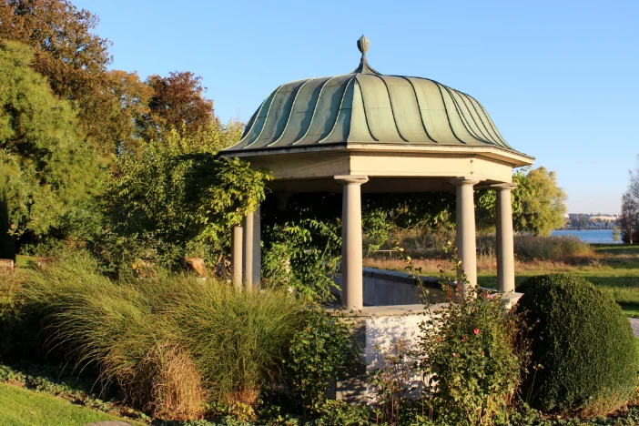 Octagonal pavilion with plain concrete column supports and a greenish rounded roof nestled among a variety of shrubbery on a clear day