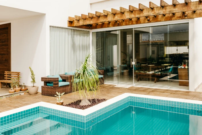 Wooden pergola attached to a white-walled house over sliding glass doors facing a deck with a swimming pool