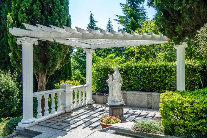 Triangular white pergola supported by columns sheltering a marble stature ona small patio surrounded by lush shrubbery and trees