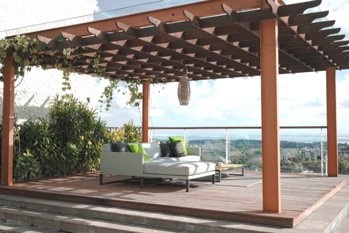 Rust pergola with vines on a square deck shading a white sofa and ottoman with bright green and taupe pillows overlooking a scenic vista