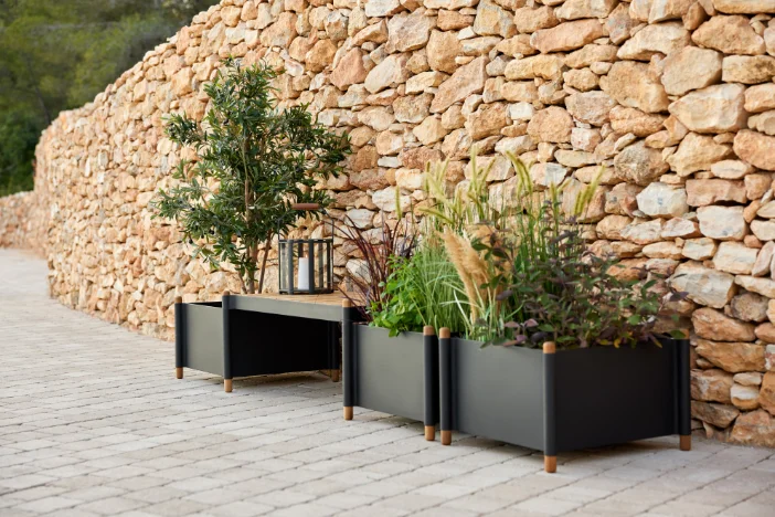 Three black square planter with greenery on a light grey brick patio in front of an orangish brown stone wall