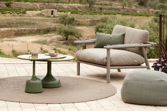 Round beige outdoor rug under two green accent tables near laid-back brown lounge chair and green pouf on a patio