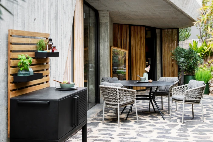 Modern dining set with round black table and woven rope chairs on a grey stone patio beneath an overhang