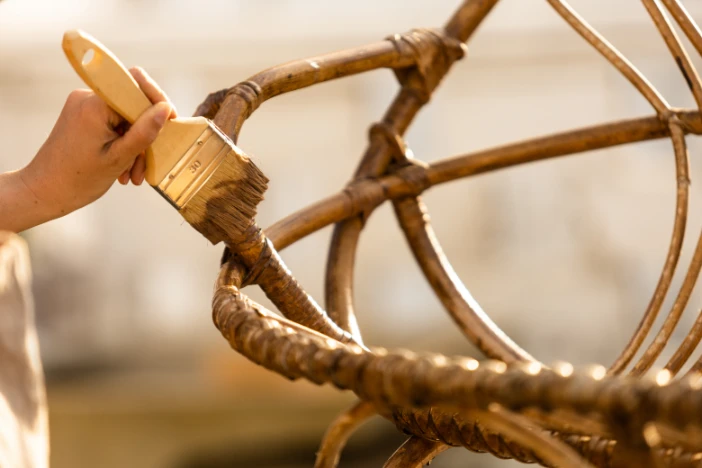 Person applying protective finish with a wide brush to a light brown rattan outdoor lounge chair