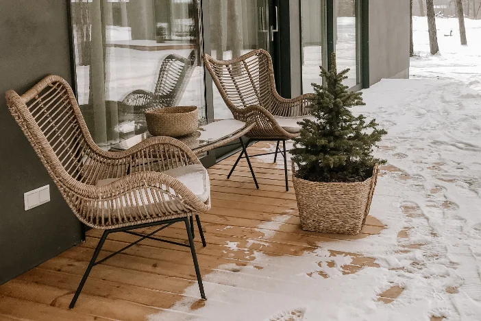 Two modern light brown wicker chairs with black metal rod legs sitting on a wooden deck with snowfall creeping up