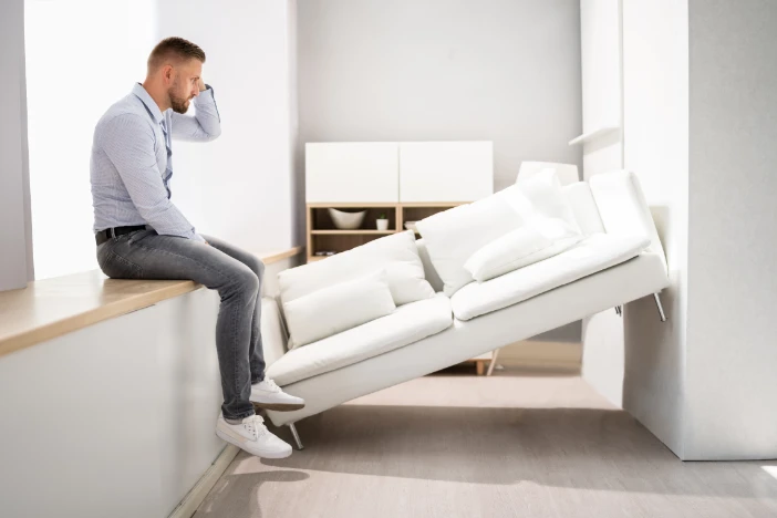 Man is sitting on a countertop near a white loveseat that is tilting in a space too small for it to set down normally