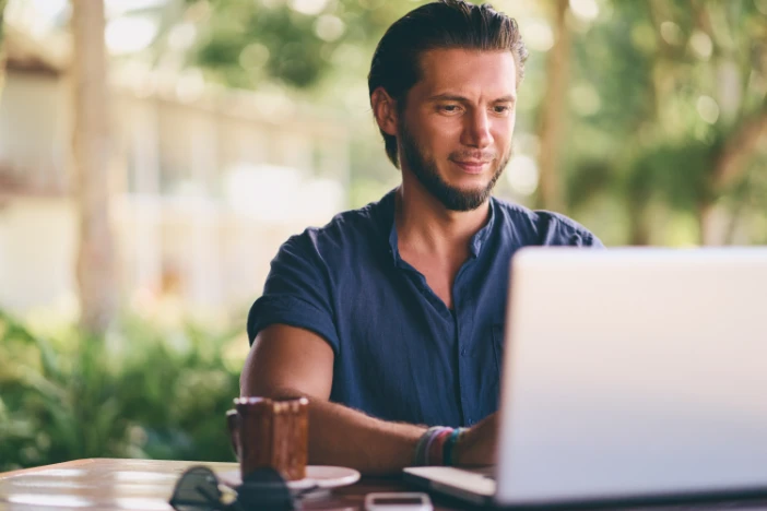 Young bearded man in blue shirt working on a laptop computer while sitting in a sunroom